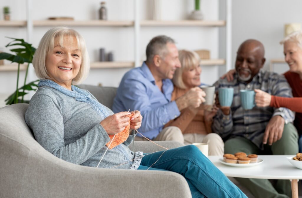 A senior sitting on a chair knitting while with friends at an assisted living community