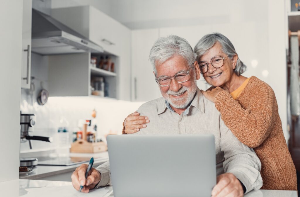 An older couple smiles while researching assisted living communities in their kitchen.