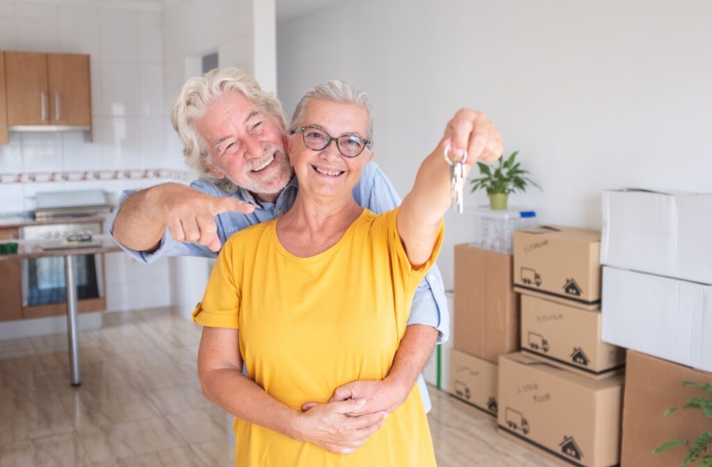An older adult stands in their packed-up kitchen with their spouse and holds up a key to their new home in assisted living