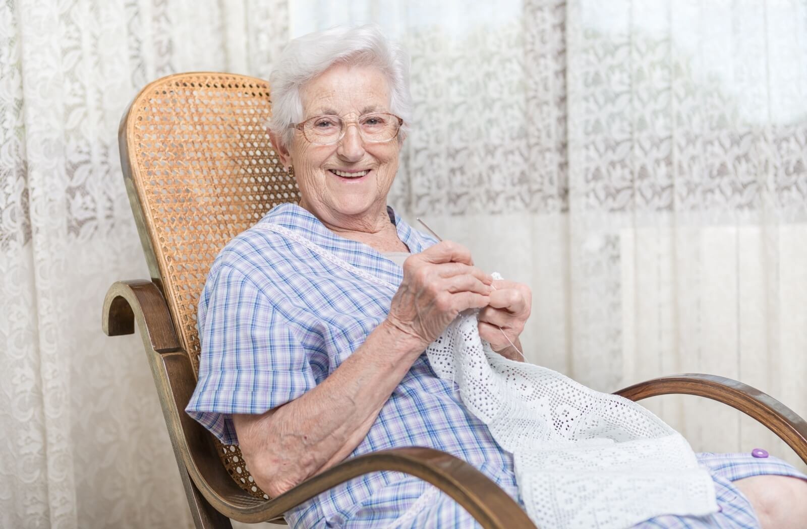 A senior smiles as they knit while sitting in a rocking chair.